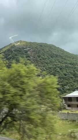 A moving shot from inside a vehicle looks out at a hilly, tree-covered landscape. In the foreground, a blurred tree with green leaves dominates the right side of the frame. Below the tree is a low-rise building with a green, sloped roof and light-colored stone walls. Another similar building is visible to the left of the first. Power lines stretch across the sky, and a tall, dark metal structure, possibly a power pylon, is visible behind the building. The sky is overcast with dramatic clouds. The camera moves slightly, revealing more of the hilly terrain and the buildings below.
