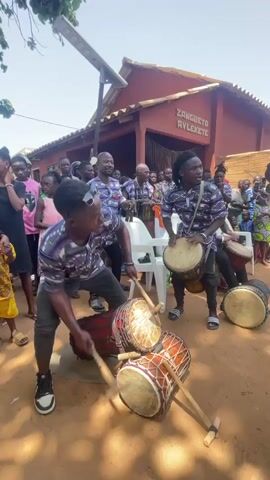 The video captures a lively outdoor scene featuring a group of people, primarily children and young adults, gathered around a building with the words "ZANGUETO AVLEXETE" visible on its facade. In the foreground, two young men are energetically playing drums. The individual on the left is crouched, rapidly striking multiple drums laid on the ground with drumsticks. The individual on the right stands, also playing a drum. In the background, more people are observing the performance, with some also holding or playing drums. The environment appears to be a dusty, outdoor area with a building and trees visible. The overall atmosphere suggests a cultural or celebratory event.