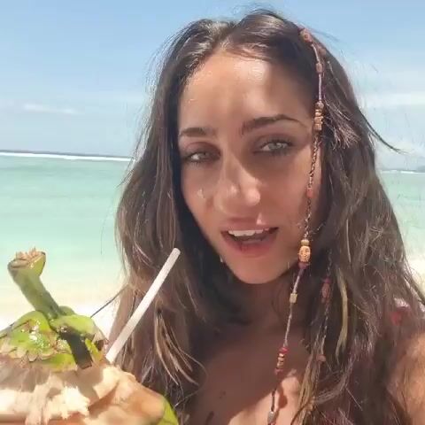 A young woman stands on a sunny beach holding a coconut with a straw. The coconut is mostly white in the middle, with green outer skin and a green stem. She looks into the camera, smiles, and moves the coconut. She has long brown hair, with beaded strands woven into her hair. The beach has white sand and the water is a clear, light blue.