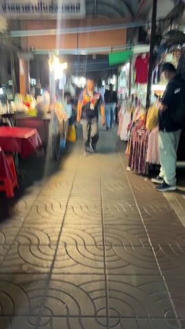 The video shows a narrow street market scene. Several people are walking through the market, which is lined with stalls selling food and clothing. Some people are eating at a table on the side of the street. The lighting is dim, and the stalls are lit by overhead lights. The ground is paved with a pattern of interlocking circles. The street is crowded with people and merchandise.