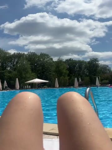 The video opens with a close-up shot of a person's bare legs and knees, which are wet, indicating they are likely near or in a pool. The camera then pans slightly to reveal a large, bright blue swimming pool under a partially cloudy sky. Several white beach umbrellas are set up around the pool, and lounge chairs are visible in the background. Trees form a dense green line behind the pool area. A pink inflatable object, possibly a flamingo float, is floating in the pool. A metal ladder with a silver handle is partially submerged in the water, near the edge of the pool. The scene is outdoors and appears to be a sunny day at a resort or a public pool.