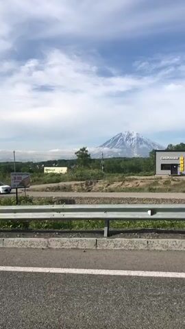 The video shows a distant snow-capped mountain under a cloudy sky. In the foreground, a white vehicle is driving on a road next to a metal guardrail. A lamppost stands to the left of the frame, partially obscuring the view of the road. There are trees and some buildings in the mid-ground.