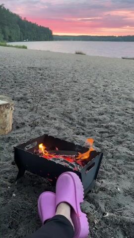 The video opens with a close-up of a small, black, metal fire pit on a sandy beach. Flames and glowing embers are visible within the pit, indicating an active fire. In the foreground, a pair of bright pink Crocs are visible, suggesting someone is sitting in front of the fire pit. The camera then pans upward, revealing a wide expanse of sandy beach leading to a calm body of water. In the distance, a tree-lined shore is visible, and the sky is filled with a dramatic sunset, featuring vibrant pinks, oranges, and purples, with some darker clouds adding depth to the scene. The overall impression is a peaceful, serene setting at dusk.
