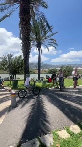 The video opens with a child in a white helmet riding a green bicycle on a paved path lined with palm trees.  A woman in patterned pants and a white top stands nearby. In the background, a body of water is visible with people sitting on benches along the shore and others walking around. The camera follows a person walking under a stone pergola with a slatted roof, casting shadows on the ground. A group of people, including children, walks through the pergola. The scene shows a grassy area with palm trees and other people enjoying the outdoor space.