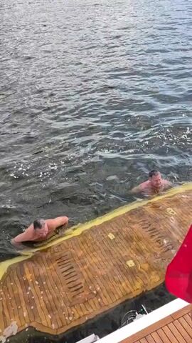 Two men are in the water next to a wooden structure. One man, wearing blue swim trunks, is on his stomach, holding onto the edge of the wooden structure with his arms and kicking his legs. The other man, to the right and further back in the water, is treading water and looking towards the man in the swim trunks. The wooden structure appears to be part of a boat or dock, with a yellow edge. The water is dark and choppy.