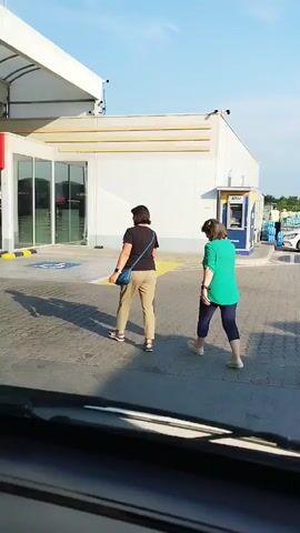 Two women walk towards a building with a red sign that says "VIVA". The woman on the left wears a dark shirt and tan pants, carrying a blue purse. The woman on the right wears a green shirt and dark pants and carries a large yellow object. They are walking on a paved area with yellow and blue bollards. The background shows a modern building with large windows and a covered area, possibly a gas station or convenience store. The sky is blue and clear. The view is from inside a vehicle looking out through the windshield.