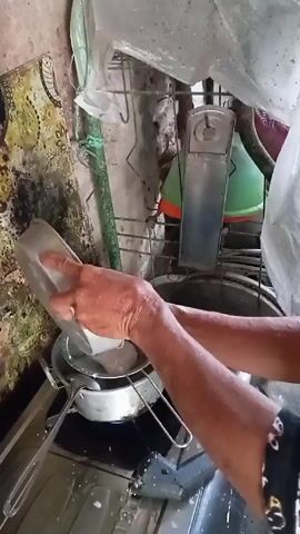 Here's a detailed description of the video:

A person is shown squeezing grated coconut in what appears to be a kitchen or food preparation area. The person is using their hands to squeeze the grated coconut over a metal strainer, collecting the liquid. The coconut and strainer are held over a metal pot. The surrounding environment looks somewhat cluttered and aged, with various kitchen utensils and cleaning supplies visible. Some of the wall areas show signs of wear and possible mold or staining. The lighting is somewhat dim, which adds to the slightly unhygienic appearance of the surroundings. The actions suggest the person is extracting coconut milk from the grated coconut.