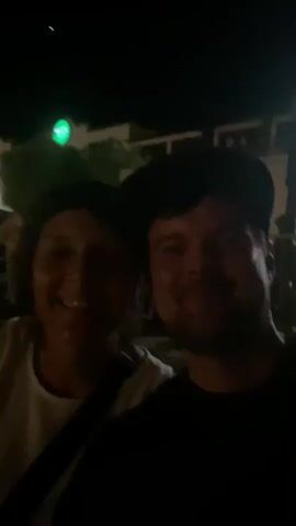 The video captures a nighttime scene in Tavira, Portugal. The initial frames show a selfie of two individuals, a woman and a man, smiling at the camera. The woman is wearing a white shirt, and the man is wearing a dark t-shirt and a black baseball cap. Behind them, a building with a green circular sign, possibly a shop, is visible. The lighting is dim, suggesting it is dark outside.

As the video progresses, the camera pans away from the selfie to show a view of a river or body of water. In the distance, a bridge is illuminated with lights, reflecting on the water's surface. The lights create a blurred, streaky effect due to the movement of the camera and the low light conditions. The overall atmosphere appears to be festive or recreational, possibly during an event or an evening out. The time of day is clearly night, and the weather appears to be clear, as there are no indications of rain or adverse conditions. The location cues, such as the bridge and the general ambiance, are consistent with a European city. The emotions conveyed by the individuals in the selfie are happiness and enjoyment. No legible text is clearly discernible, apart from the blurry green sign in the background.