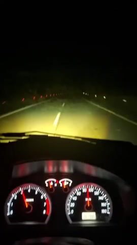 The video shows a first-person perspective from inside a car driving on a road at night. The car's dashboard is prominent in the foreground, with the speedometer on the right indicating a speed of approximately 100 units (likely km/h or mph) and the tachometer on the left showing an engine speed of roughly 3000-4000 RPM. The odometer displays "167395". The road ahead is illuminated by the car's headlights, revealing white lane markings and distant red lights on the left side of the road. The surroundings are dark, indicating nighttime driving.
