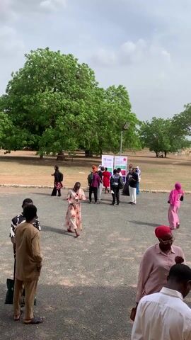 The video shows a group of people, mostly dressed in traditional attire, gathered in an open outdoor space. The area appears to be a courtyard or a large parking area next to a park with several trees. Some people are standing and chatting, while others are walking. There is a sign with the logo "Awibi HEALTHCARE" and some text in front of them. A light post is seen in the background. The weather appears to be overcast. The people are diverse in their clothing and ages.