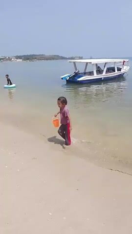 The video shows a beach scene on a sunny day with clear blue skies. In the foreground, a young girl with dark hair and wearing a pink shirt and gray pants walks along the sandy shore, carrying an orange bucket. She is walking towards the right side of the frame. Further back, on the beach and in the shallow water, several other children are playing. One child is sitting in the sand, seemingly building something, next to a yellow inflatable duck. Another child is in the water with a pink inflatable ring. A white boat with a blue trim is anchored in the calm, clear water. Along the shoreline in the background, there are several temporary structures, possibly stalls or awnings, and trees. The overall atmosphere appears to be one of leisure and recreation.