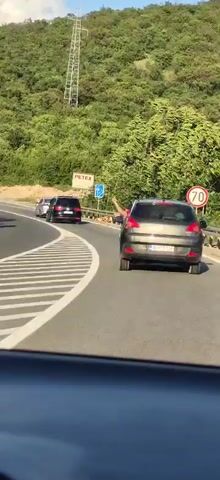 The video shows a car driving on a highway behind two other cars. The car in front of the vehicle recording the video is gray. The car in front of that is black. Both vehicles are traveling on a paved road with a white line separating their lane. There is a wooded hillside to the right and left of the road. The driver of the gray car appears to be holding up a flag out the window, and it waves in the wind. Signage indicating 'PETEX' is visible on the side of the road.