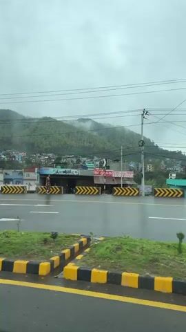 The video captures a street scene in Vyas (Damauli), Nepal, viewed from a moving vehicle on an overcast day with recent rain. The road is wet, and green, mist-covered hills are visible in the background. Multi-story buildings, some residential and some under construction, line the roadside.

A central median with grass and distinctive yellow and black concrete blocks separates the road lanes. Traffic includes a brown and orange bus with a green, white, and red stripe, and two motorcyclists, one wearing a yellow raincoat. Later, a white, blue, and red bus passes, and pedestrians, some holding umbrellas, are seen on the sidewalk. Electrical poles and wires are visible overhead. Faint Nepali text appears on storefronts, including "जिन्दगि" (Zindagī, meaning "life") and what seems to be a shop name, "सिष्ट फूट पसरा".