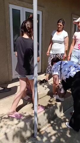 FIVE PEOPLE STANDING OUTSIDE A BUILDING. TWO GIRLS IN THE FOREGROUND ARE FACING AWAY FROM THE CAMERA AND TOWARDS THREE WOMEN WHO ARE FACING THE CAMERA. THE TWO GIRLS ARE WEARING SHORTS AND SANDALS. THE WOMAN ON THE FAR LEFT IS WEARING A BLACK TANK TOP AND RED SHORTS. THE WOMAN NEXT TO HER IS WEARING A BLACK T-SHIRT AND DENIM SHORTS. THE WOMAN IN THE MIDDLE IS WEARING A WHITE T-SHIRT AND FLORAL PANTS. THE WOMAN ON THE FAR RIGHT IS WEARING A PINK T-SHIRT AND BLUE SHORTS. A WOMAN IN THE FOREGROUND IS BENDING DOWN TO ADJUST SOMETHING ON HER FEET. THE BUILDING HAS A LIGHT COLORED EXTERIOR AND A GLASS DOOR. THERE IS ALSO A METAL POLE IN THE FOREGROUND. THE GROUND IS COVERED IN CONCRETE AND SHADOWS FROM TREES.