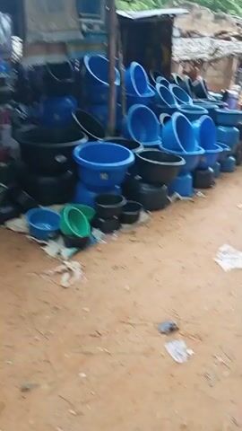 A street market stall displays numerous plastic basins in various sizes and colors, predominantly blue and black, with some green ones in the foreground. The basins are stacked and arranged in rows. Behind the basins, there is a stall structure with a corrugated metal roof and wooden supports. In the background, more stalls are visible, along with people moving around. The ground is sandy and dusty, with some debris scattered around. The overall impression is a busy outdoor market scene.