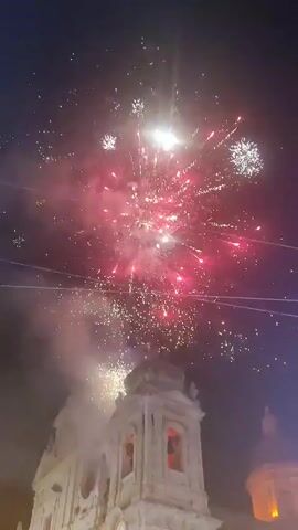 The video shows a nighttime fireworks display above a white building with a dome. The fireworks are various colors, including white, gold, and red. Smoke from the fireworks obscures parts of the building. The video appears to be shot from a low angle, looking up at the fireworks and the building. There are power lines visible in the foreground.