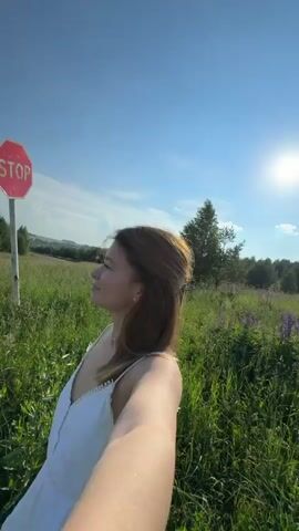 A young woman with shoulder-length auburn hair, wearing a white dress and a bracelet, is filmed in a selfie-style video in an outdoor setting. She is standing in a field of tall green grass under a clear blue sky with scattered clouds. In the background, a stop sign is visible on a pole, along with trees and a dirt road. The woman turns her head, looks at the camera, and smiles. The camera angle shifts, showing her from above as she looks down and smiles again.