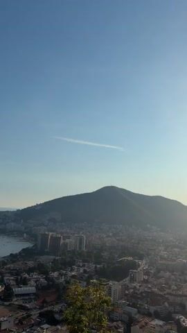 The video shows an aerial view of a coastal city and the sea. The camera pans to the left, revealing a bay with numerous boats docked. The city, nestled between a large hill and the sea, features a mix of modern buildings and older structures. A clear blue sky dominates the upper portion of the frame. Greenery, including trees and shrubs, is visible in the foreground and on the hillsides. The overall impression is a scenic vista of a seaside town bathed in daylight.
