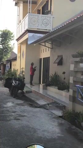 A person wearing a red shirt, gray pants, and a tan cap stands barefoot in front of a house. They are holding a mobile phone and appear to be looking at it. A black motorcycle is parked on the left side of the image. The house has a tan exterior with dark-colored double doors. The scene appears to be outdoors, possibly on a street or driveway.