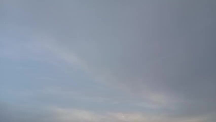 The video shows a cloudy sky with some wispy clouds visible. The light suggests it is either dawn or dusk, as the sky is overcast and lacks direct sunlight. The bottom left corner of the frame shows a small portion of what appears to be a dark structure, possibly a building or a silhouette of a distant object. The camera is mostly focused on the sky, which is predominantly gray and light blue, with some areas appearing darker, indicating thicker cloud cover. There are no people or clear objects visible in the frame, and no discernible actions or interactions are taking place. The overall impression is a natural outdoor scene with overcast weather.