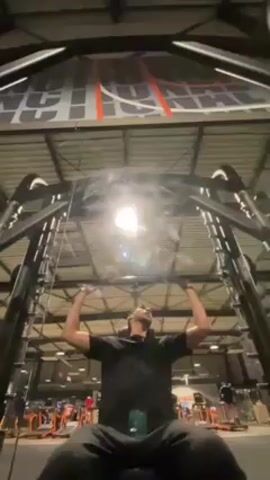 A man is seated in a weight training machine in what appears to be a gym. The man is wearing a black hoodie and black sweatpants. He is positioned beneath the bar of the machine, with his hands gripping the bar above his head. He performs a lifting motion, extending his arms upwards. The gym has a high ceiling with exposed metal beams and a sign in the background. Other exercise machines and people can be seen in the periphery.