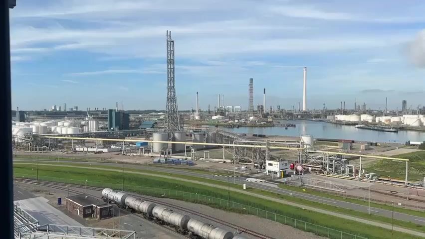 This is an aerial drone video capturing a sprawling industrial complex likely involved in oil refining or petrochemical production. The scene features a large body of water, possibly a harbor or canal, with several ships docked, including a prominent red tanker. Extensive infrastructure is visible, including numerous large white storage tanks, tall industrial chimneys or flare stacks emitting steam or smoke, a network of pipes and scaffolding, and various buildings and processing units. In the foreground, a set of train tracks runs parallel to a road, with several large, cylindrical tank cars on the tracks. The environment is industrial, with a clear sky and some scattered clouds, suggesting daytime. The camera pans slowly across the landscape, offering a broad overview of the facility. There are no visible people in the footage.