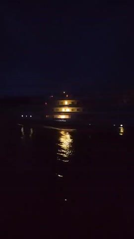 The video shows a three-deck vessel moving on water at night. The vessel has lights on, and their reflections are visible in the water. The sky is dark, suggesting it is nighttime. The vessel moves across the frame from left to right, leaving ripples in the water.