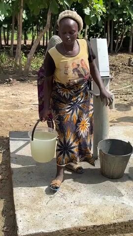 A woman wearing a light-colored beanie, a tan tank top with an image of two women, and a dark, patterned skirt walks towards a water pump. She is carrying a light-yellow bucket in her left hand. A young child carrying another child on their back is visible in the background to her right. Several men and another child are standing in a group to her left, observing her. The ground is dirt and some concrete. There are trees and vegetation in the background, as well as a simple building. The woman approaches the pump, and the video continues to show her from behind as she bends down. The bucket is placed on the ground near the pump.