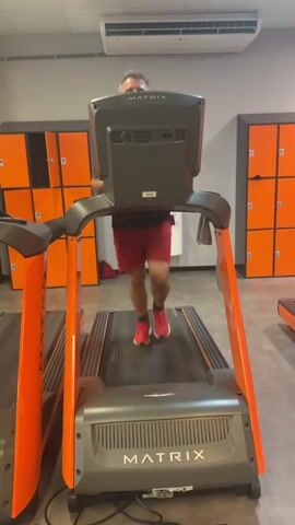 A man is running on a treadmill in a gym. He is wearing red shorts and pink athletic shoes. The treadmill is orange and black and has the brand name "MATRIX" on it. In the background, there are orange lockers. The man is smiling at the camera.