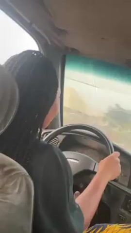 A person with long dark braids is driving a car. The driver is wearing a dark shirt and patterned pants. The car appears to be old, with a crack in the windshield. The scenery outside the car window is blurry, but appears to be a landscape with vegetation. The car's interior is worn.