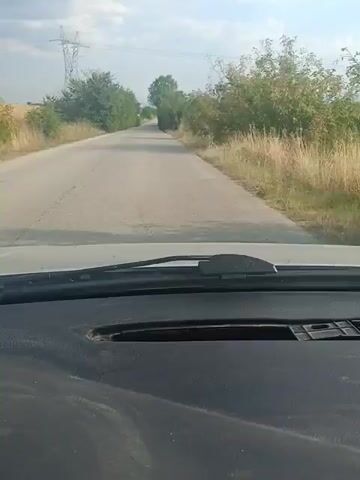 The video shows a first-person perspective from inside a vehicle, looking out through the windshield. The road ahead is paved and appears to be a rural or country road, with a slight curve to the right. The road is flanked by vegetation on both sides, consisting of trees and tall dry grass. In the distance, a power line pylon can be seen on the left side of the road. The sky is partly cloudy. The dashboard of the vehicle is visible in the foreground, showing a dark, textured surface with a rectangular indentation. The windshield wipers are positioned at the bottom of the windshield. The video captures the movement of the vehicle as it travels down the road.