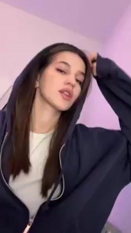 A young woman with light skin and dark brown hair is positioned in the frame, wearing a navy blue hooded zip-up sweatshirt over a white top. The hood is up, framing her face. She has eyeliner on her upper eyelids, and her lips are slightly pursed. She glances directly at the camera, offering a small smile. She has a small stud earring in her ear, along with a ring ear cuff. The background is a light pink-purple gradient with a hint of white in the upper left corner. The camera angle is slightly tilted, providing a close-up view of her face. The lighting is soft, enhancing her features.
