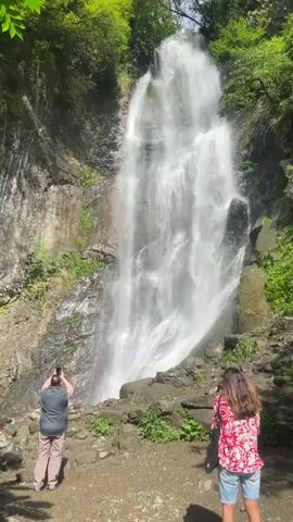 A man in a gray shirt and khaki pants stands on the left, holding a phone to his face, seemingly taking a picture of a tall waterfall. A woman in a red and white patterned shirt and denim shorts stands to the right, facing the waterfall. Behind her, a man in a maroon shirt walks to the right. Two other people, one in a white shirt and the other partially obscured, are on the far left, walking towards the waterfall. The waterfall cascades down a rocky cliff face surrounded by lush green trees and vegetation. The ground in front of the waterfall is a dirt and gravel path.