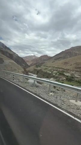 A first-person perspective video shows a view from a moving vehicle, likely a car, on a paved road. The road runs alongside a steep, rocky mountain slope. A metal guardrail separates the road from the slope. In the valley below, a river flows through a sparsely vegetated area. There are a few small buildings clustered together in the distance. The sky is cloudy with patches of blue visible. The overall environment appears to be a mountainous, arid region.