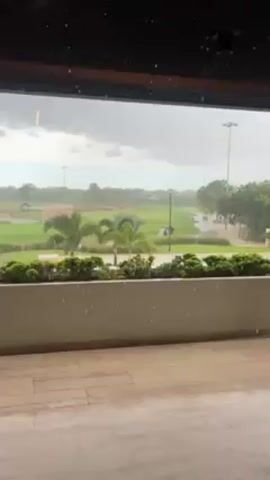 The video depicts a view through a window onto a golf course during heavy rain. The foreground consists of a tiled porch or balcony, with a low wall topped with greenery. Beyond this, the golf course stretches out, with green fairways, sparse trees, and a few water features. A golf cart is driving along a path. The sky is overcast and gray, contributing to the overall feeling of a gloomy, rainy day. Raindrops are visible on the window, which is framed by a dark structure, possibly the roof or a beam of the building.