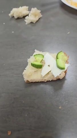 The video displays a static, close-up shot of a small, open-faced snack resting on a grey, textured surface, likely a tabletop or counter in an indoor setting.

The main subject is an irregularly shaped piece of white bread, approximately square, with a visible crust edge on its right side. This bread is topped with several thin, irregular pieces of white cheese and four quarter-slices of green cucumber. The cucumber pieces are arranged in two pairs, with one pair at the top and another at the bottom of the bread, flanking the cheese.

In the background, two smaller, torn pieces of white bread are visible to the left. On the far right, a portion of an orange utensil handle (possibly a fork) and a white plate containing some yellow residue (resembling cooked egg) are partially visible. No people are present, and no specific activity is shown beyond the presentation of the prepared snack. There is no visible text in the frame. The time of day and weather are not discernible. No specific location cues beyond an indoor setting are present within the video itself.