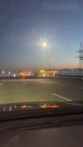 This media file captures a view from inside a vehicle driving on a highway at dusk. The sky is a gradient of deep blue, transitioning to lighter shades of blue and hints of pink and orange near the horizon, suggesting the sun has recently set or is about to rise. A bright streetlamp illuminates the road ahead. In the distance, a gas station with a yellow canopy and illuminated signs stands out. The gas station appears to be modern and well-lit, with a few vehicles visible. The overall scene is serene, with a sense of transition, possibly a journey or a late arrival. The headlights of the car are on, casting a glow on the road. The dashboard of the car is partially visible in the foreground. The weather appears clear, with no significant precipitation. The overall mood is one of quiet travel as nightfall approaches.