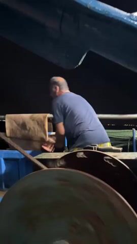 A person wearing a dark blue t-shirt and dark pants is on a boat. They are holding a long pole with a cloth at the end and are using it to push a large, brown, rectangular object. The boat has blue and dark metal structures visible, and what appears to be a large winch or spool in the foreground. The background is dark, suggesting it is nighttime or a poorly lit area. The person's actions suggest they are cleaning or adjusting something on the boat.