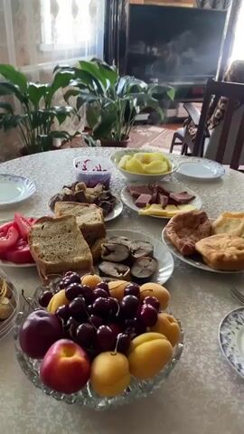 The video displays a table laden with food, suggesting a meal or a spread of refreshments. Various dishes are visible, including sliced bread, sliced tomatoes, cooked potatoes, and what appears to be cured meats and cheese. There is also a bowl of fruit, containing cherries, apricots, and apples, and a container of chocolate candies. In the background, potted plants are placed near a window, and a television set is visible in the distance. The setting appears to be an indoor environment, possibly a dining room or living area. No people are visible in the frame.