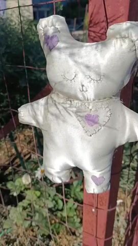 This video captures a charming garden scene, likely in Kuchugury, Russia, on a bright, sunny day. The primary subject is a well-loved, white stuffed teddy bear, adorned with small purple hearts. It's attached to a rusty red metal fence, its fabric shimmering in the sunlight. The bear appears to be a cherished toy, perhaps left outside for a moment.

The fence itself is a significant element, showing signs of age with its rust and the wire mesh. Beyond the fence, a lush garden unfolds. We see vibrant green bushes, likely rosemary, forming a dense backdrop. Scattered amongst them are delicate white daisies with yellow centers, adding pops of color. Further back, rows of lavender bushes are visible, their purple hues creating a soothing contrast. The overall impression is one of a well-tended, peaceful outdoor space.

The camera pans across the garden, revealing more of its beauty. The rough texture of gravel paths and stone borders is evident, suggesting a natural and rustic aesthetic. In the distance, hints of buildings and trees suggest a residential area, reinforcing the location in Kuchugury. The sunlight casts dappled shadows, indicating it's likely mid-morning or afternoon. There are no people visible in the video, and no text is present. The overall mood is serene and bucolic, with the slightly worn teddy bear adding a touch of nostalgic sweetness to the scene.