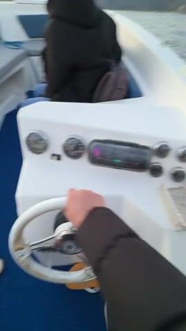 The video shows the interior of a small boat as it moves across water. A person wearing jeans and a dark jacket is seated on a bench in the boat. Someone wearing white shoes is at the helm, operating the steering wheel. The boat is moving towards a sunset on the horizon, with land visible in the distance. The boat has blue carpeting and white sides with cushioned seating. There is an instrument panel with gauges on the steering console. The camera shakes, indicating movement. Music plays in the background.
