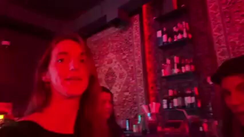 Three young women stand close together, the woman on the left films a video in what looks like a small bar, or dance club with red lighting. Behind them is a bar area with a shelf of bottles. The decor includes red patterned fabric on the walls. Music plays as the woman in the center sings. The women talk and laugh. A man stands behind the bar area.