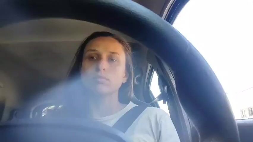 The video shows a woman in the driver's seat of a car. She is wearing a white t-shirt and a seatbelt. She looks ahead and then touches her hair. The steering wheel is in the foreground, and a street with buildings and trees is visible through the windshield. The video appears to be taken from the driver's perspective. There's a blur or reflection on the windshield.