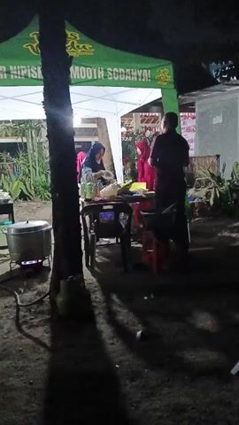 A night scene unfolds under a green canopy tent with white sides, illuminated by artificial lights. A woman wearing a hijab is visible behind a table, seemingly preparing food. A man in a dark shirt stands to her right, facing away from the camera. To the left, a large pot sits on a burner, indicating cooking activities. The ground is dark and dusty, with shadows cast by trees and the tent. The atmosphere suggests an outdoor food stall or event.