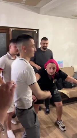 The video shows four men in a room with wooden floors and white walls. The man in the foreground on the left is wearing a white polo shirt and grey pants, and is mid-dance. To his right, a man in a pink headscarf and black t-shirt is also dancing with his arms outstretched. Behind them, another man in a white polo shirt and white shorts stands watching. In the background, a fourth man in a grey t-shirt and black pants has his hands on his head. There is a couch with bags on it to the right of the frame.