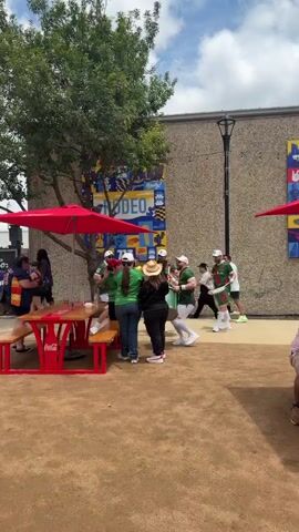 A group of five men wearing green shirts, white hats, and white shorts are playing drums outdoors. Two of the men are holding large green drums and appear to be marching. Another man in the background is also playing a drum. Two women are standing and watching the drummers. One woman is wearing a straw hat and a black top, and the other is wearing a green shirt and blue jeans. In the background, there is a mural with the word "RODEO" on it and a Coca-Cola picnic table with red umbrellas. The ground is covered in gravel.