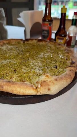The video shows a close-up of a pistachio pizza on a wooden platter, sitting on a white tablecloth. Behind the pizza are various condiments, including small bottles of hot sauce and olive oil. A pair of sunglasses is also on the table. Through the window behind the table, one can see a building with a red awning. The camera moves slightly, showing a wider view of the table.