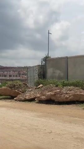 The video shows an outdoor scene under a cloudy sky. In the foreground, there is a dirt road. To the right of the road, there are large rocks and green bushes. Further back, a construction site is visible with buildings in various stages of completion, featuring concrete pillars and brickwork. A black car drives along the dirt road in the middle ground. In the background, there are hills with more construction and some trees. There is also a small, corrugated metal structure with a sign that reads "BOREHOLE DRILLING". A yellow truck is parked next to the structure.