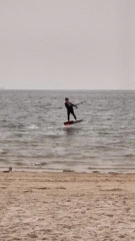 The video shows a man kiteboarding on the sea, with a person walking on the beach in the foreground. The kiteboarder is wearing a black wetsuit and uses a kite to propel themselves across the water. The board is hovering just above the water's surface, creating a hydrofoiling effect. The person on the beach walks across the screen from right to left, looking at their phone. The sky is overcast and the sea is calm. In the distance, a boat and some faint landforms can be seen. The beach is sandy.
