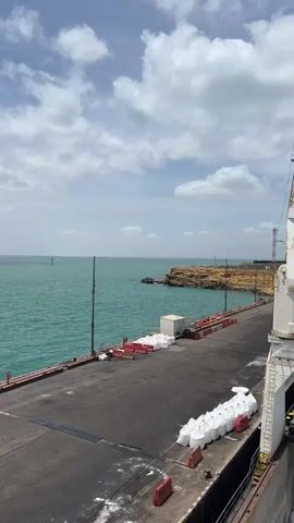 The video shows a view from a ship at a port, with a large white crane positioned over the ship's cargo hold. The hold is filled with numerous large white bags. In the background, the port's dock is visible, lined with more white bags and red and white barriers. Beyond the dock, there is a rocky, tan-colored coastline with some structures and a fence. The sea is a vibrant turquoise color under a sky with scattered white clouds. The crane is in the process of loading or unloading cargo. The scene suggests industrial activity related to shipping and bulk material handling.